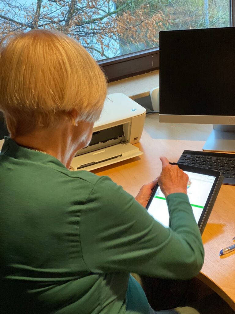 benutzerfreundliches Web Design für soziale Dienstleistungen ist wichtig für den erfolg des Unternehmens. Hier ist eine ältere Dame mit ihrem Tablet zu sehen, neben ihr steht ein Drucker und vor ihr ein Monitor.